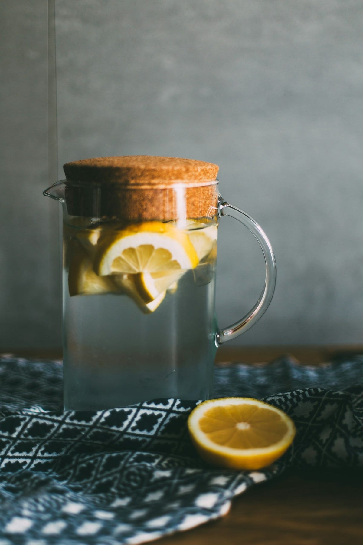 A corked glass pitcher of lemon infused water beside a halved lemon, resting on an indigo patterned cloth.
