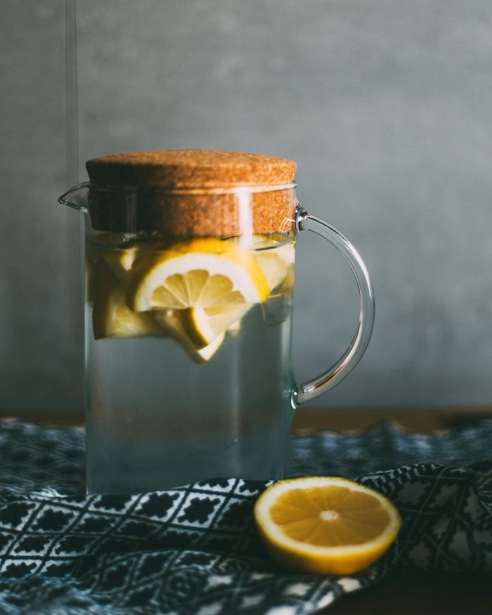 A corked glass pitcher of lemon infused water beside a halved lemon, resting on an indigo patterned cloth.
