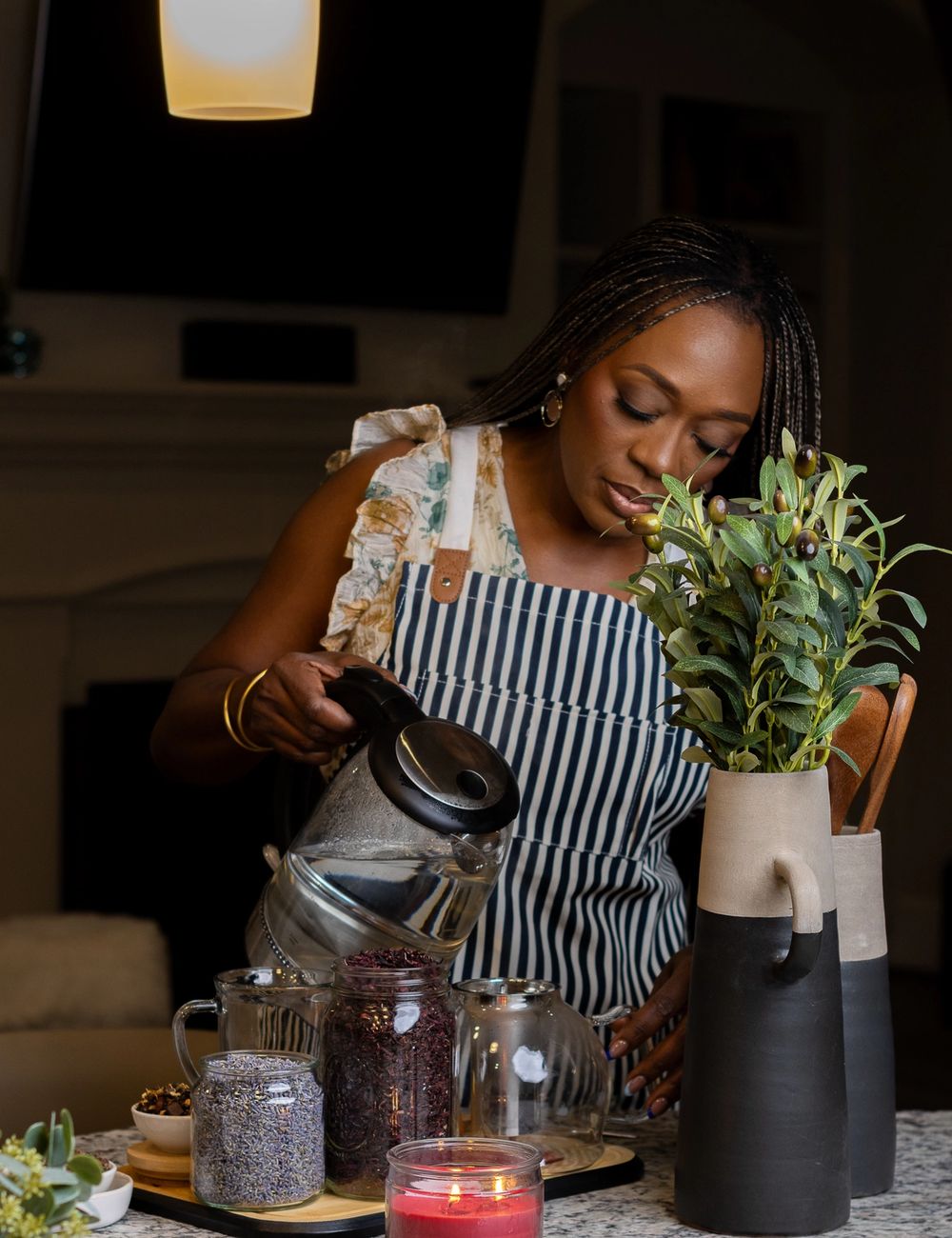Dr. Tosin tilting a black kettle to pour hot water into a clear glass teapot, apothecary jars of dried herbs and a deep-burgundy candle on the counter beside her.