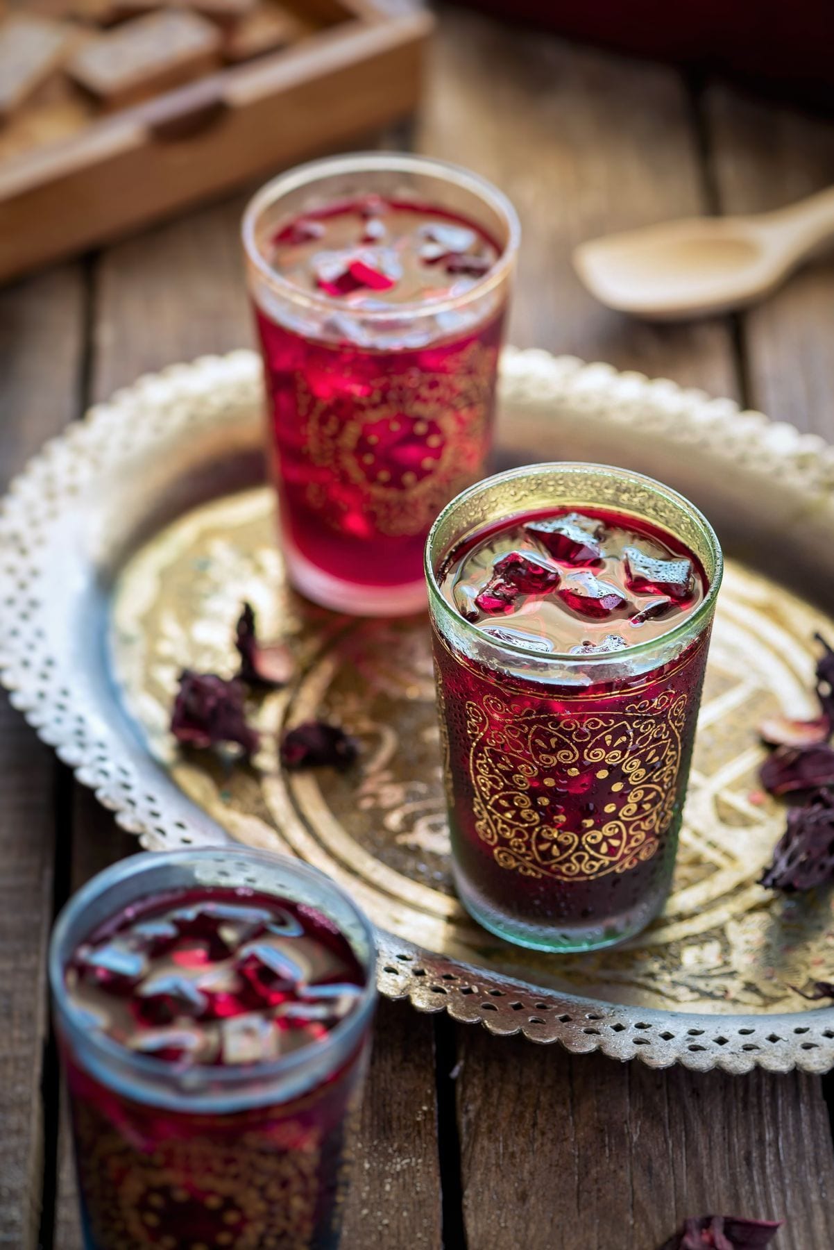 Three gold painted glasses of deep ruby hibiscus tea over ice on an ornate gold tray, dried hibiscus petals scattered beside them.