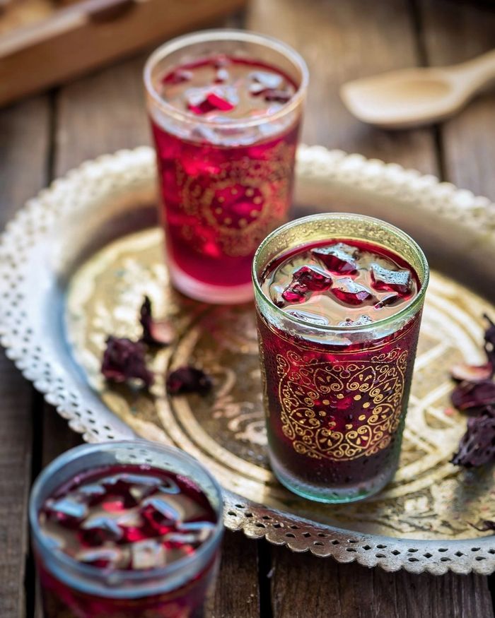 Three gold painted glasses of deep ruby hibiscus tea over ice on an ornate gold tray, dried hibiscus petals scattered beside them.