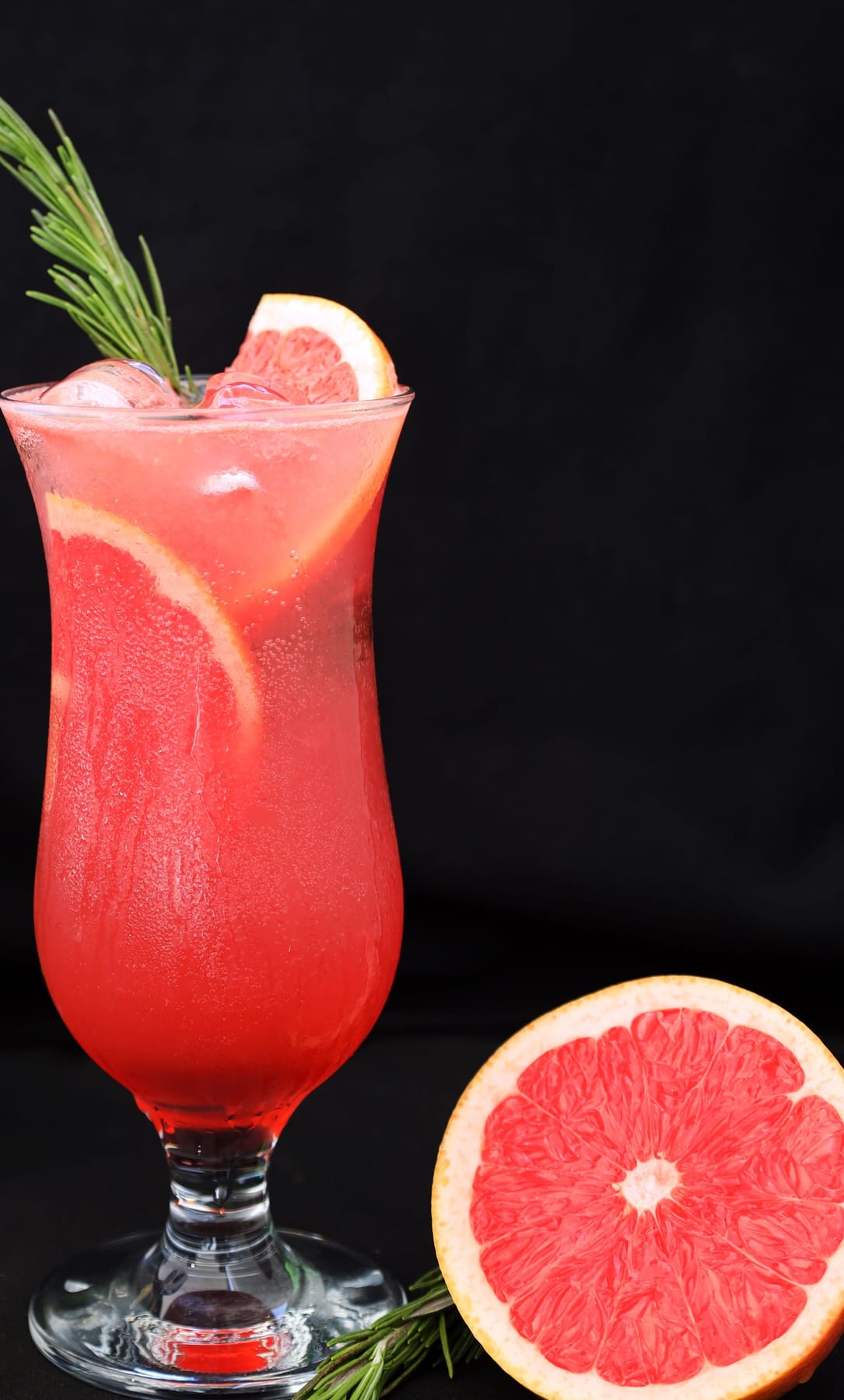 A tall hurricane glass of pink grapefruit sparkling water with a rosemary sprig and grapefruit wheel, a halved grapefruit beside it.