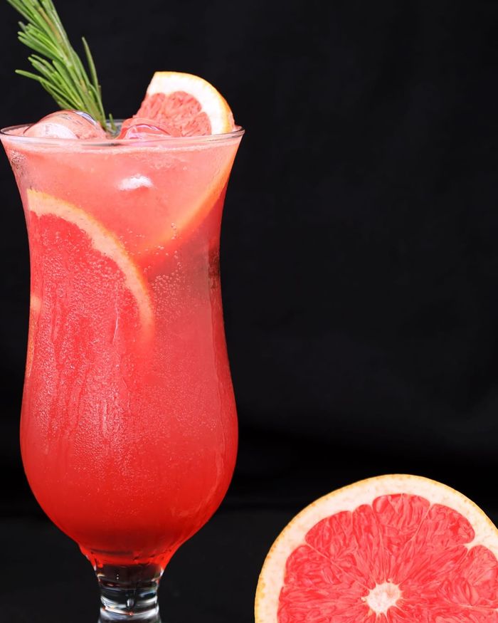 A tall hurricane glass of pink grapefruit sparkling water with a rosemary sprig and grapefruit wheel, a halved grapefruit beside it.