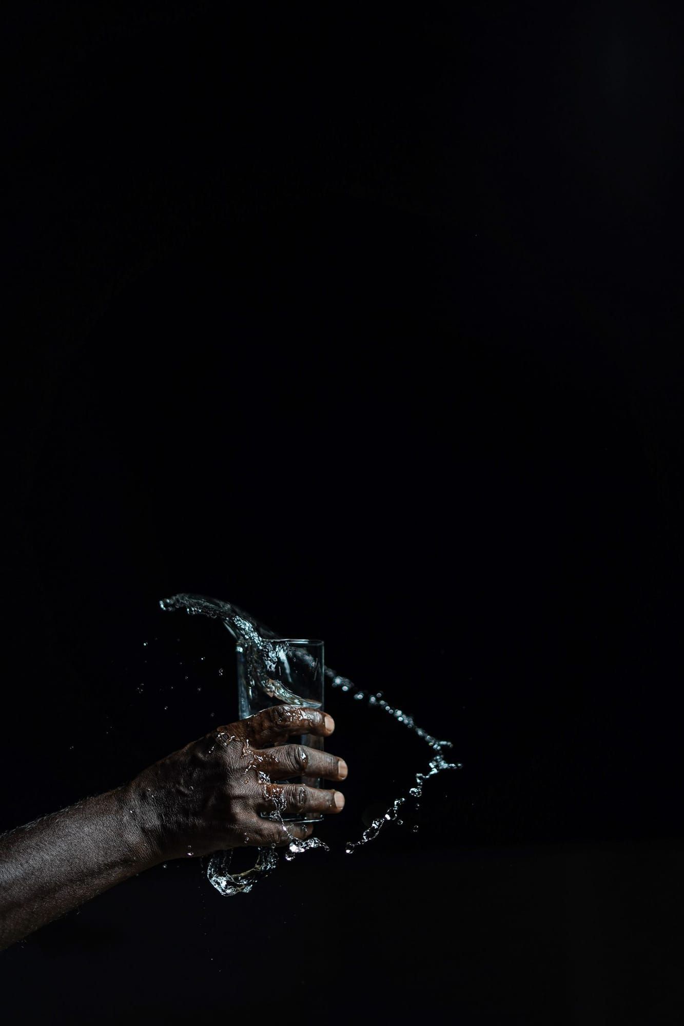 A hand holding a clear glass of water against a dark background, with water splashing around the glass.
