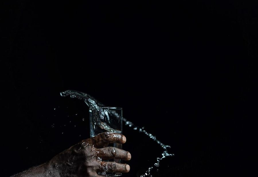 A hand holding a clear glass of water against a dark background, with water splashing around the glass.