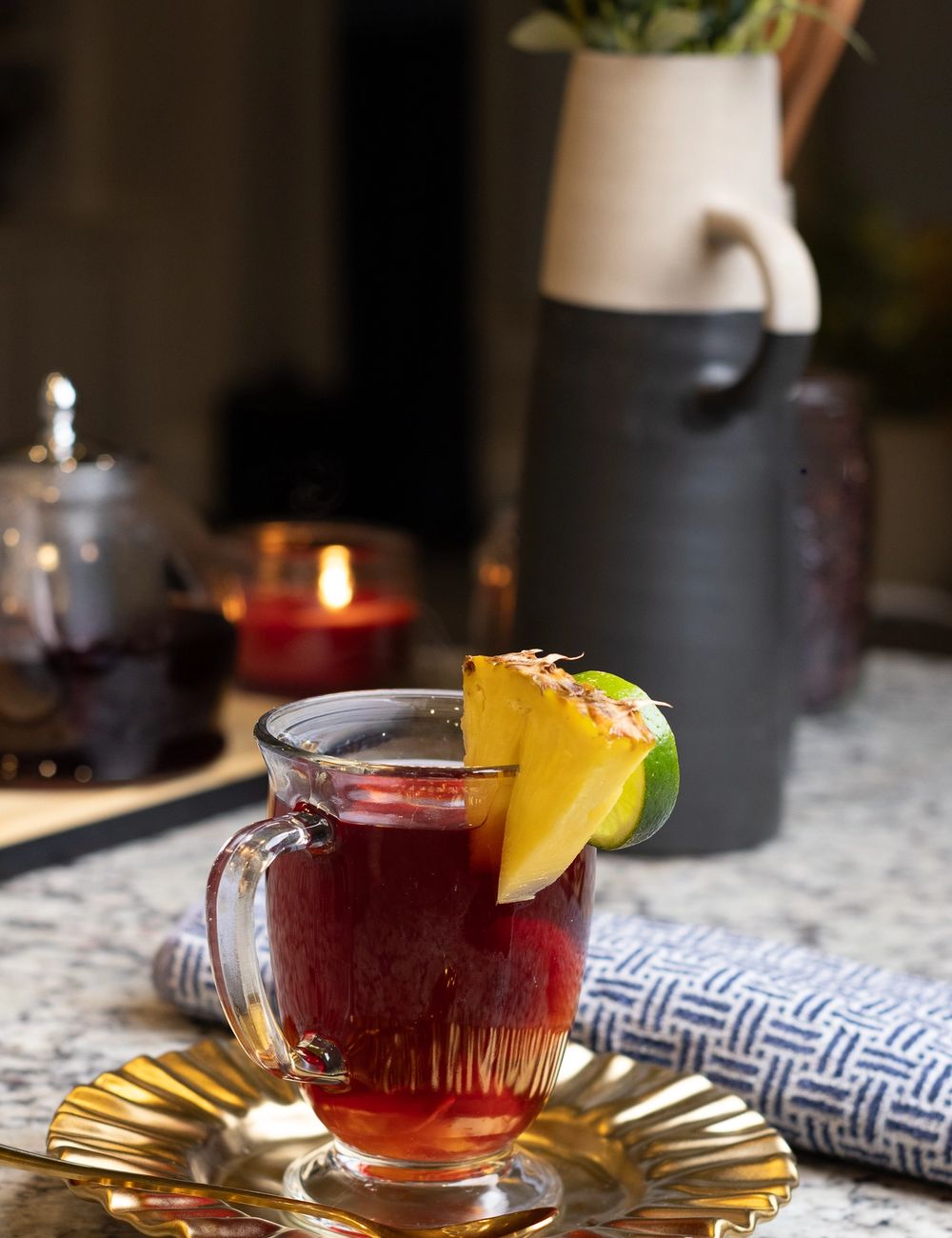 Faceted clear mug of deep ruby hibiscus tea with a citrus wedge garnish, perched on a gold scalloped saucer beside a small carafe and folded indigo-stripe napkin.