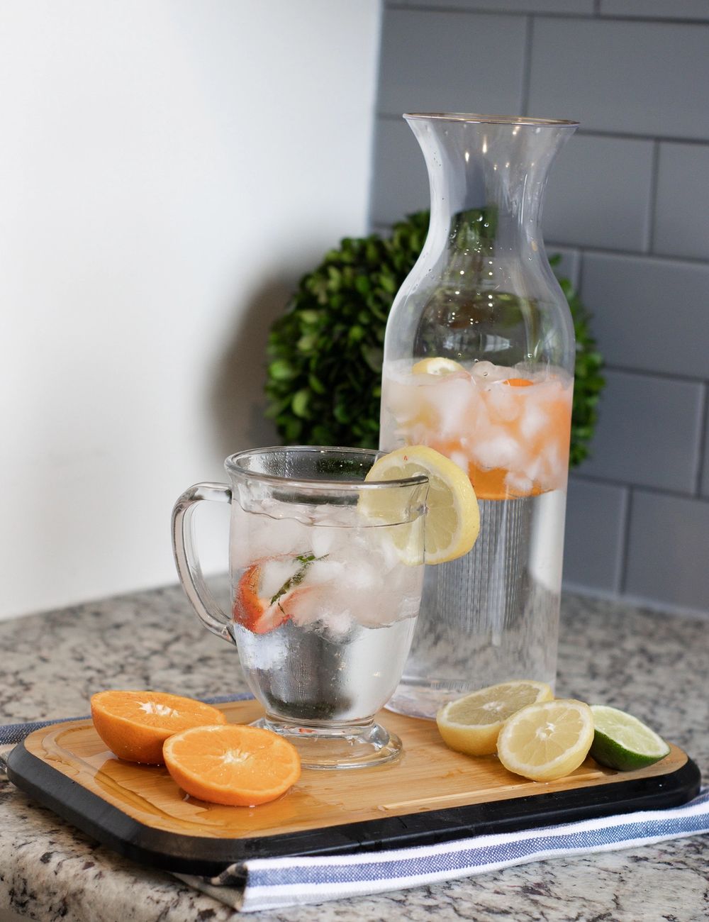 A glass carafe and matching mug of soft-pink fruit-infused water, strawberry and citrus slices floating, kitchen counter beside a green sprig in a textured planter.