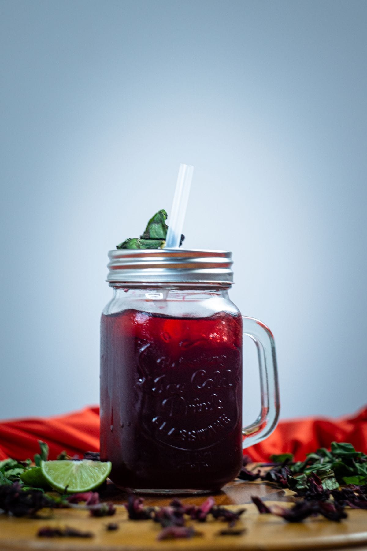 A mason jar mug of deep ruby hibiscus cooler with a lime wedge at its base, dried hibiscus petals scattered on the wood around it.