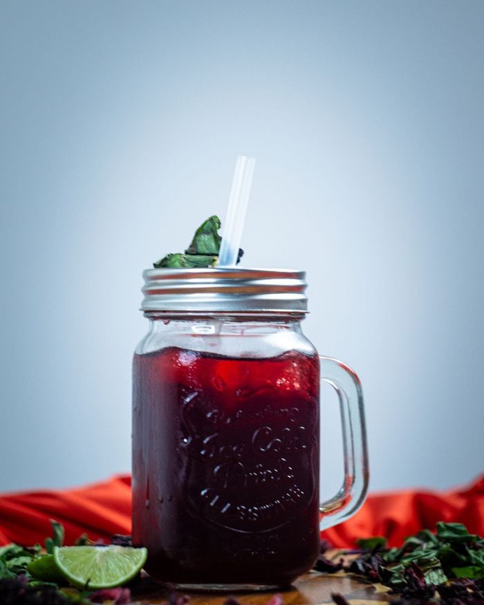 A mason jar mug of deep ruby hibiscus cooler with a lime wedge at its base, dried hibiscus petals scattered on the wood around it.