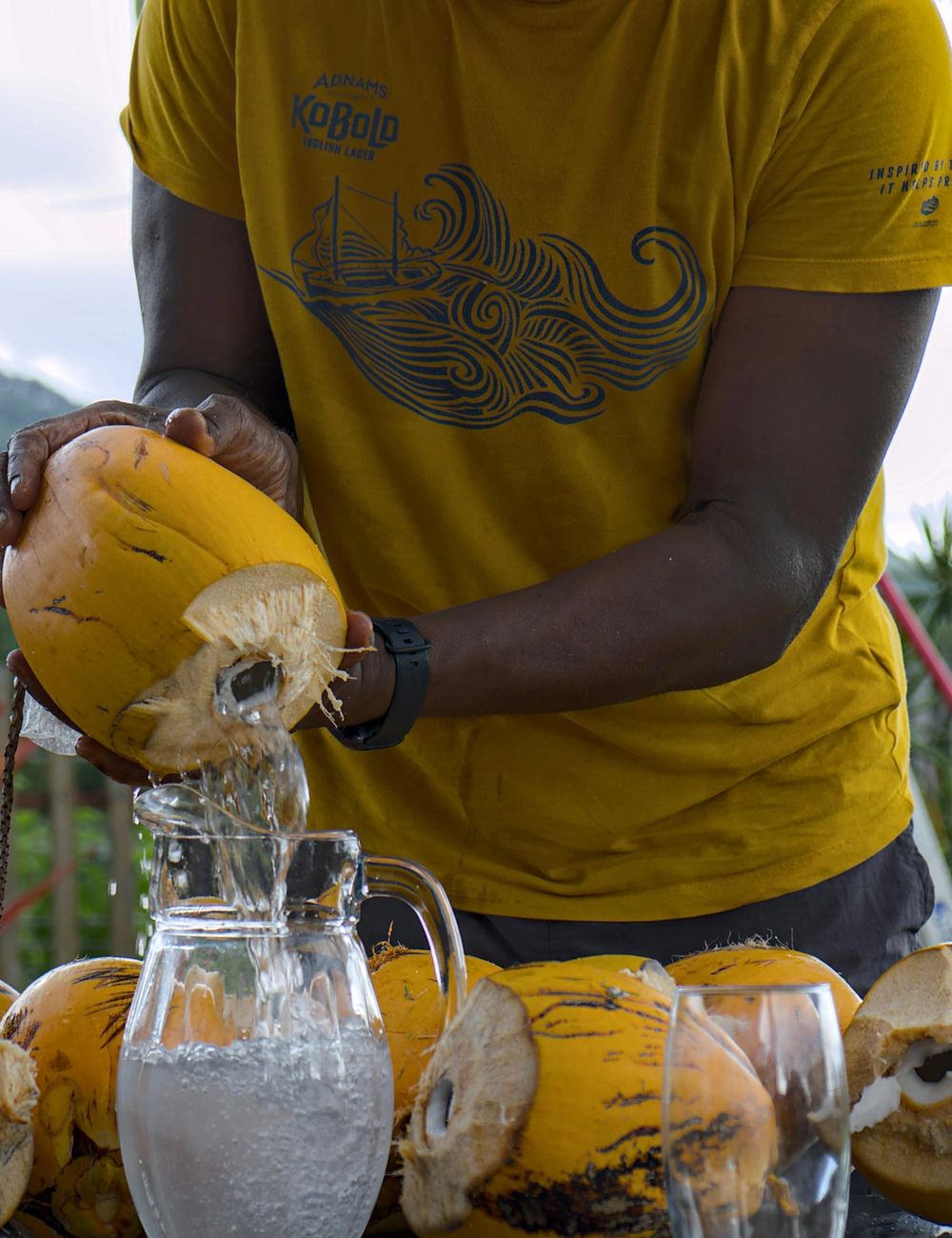 Black hands pouring fresh coconut water from a halved yellow coconut into a clear glass jug, more whole coconuts crowded on the table behind, soft daylight outdoors in Trinidad and Tobago.
