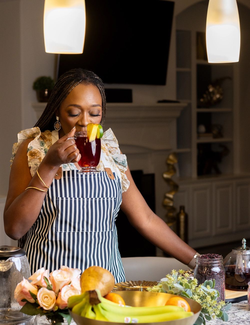 Dr. Tosin sipping from a faceted tea mug at a marble kitchen island, fresh fruit bowl and cookbooks softly out of focus behind her.