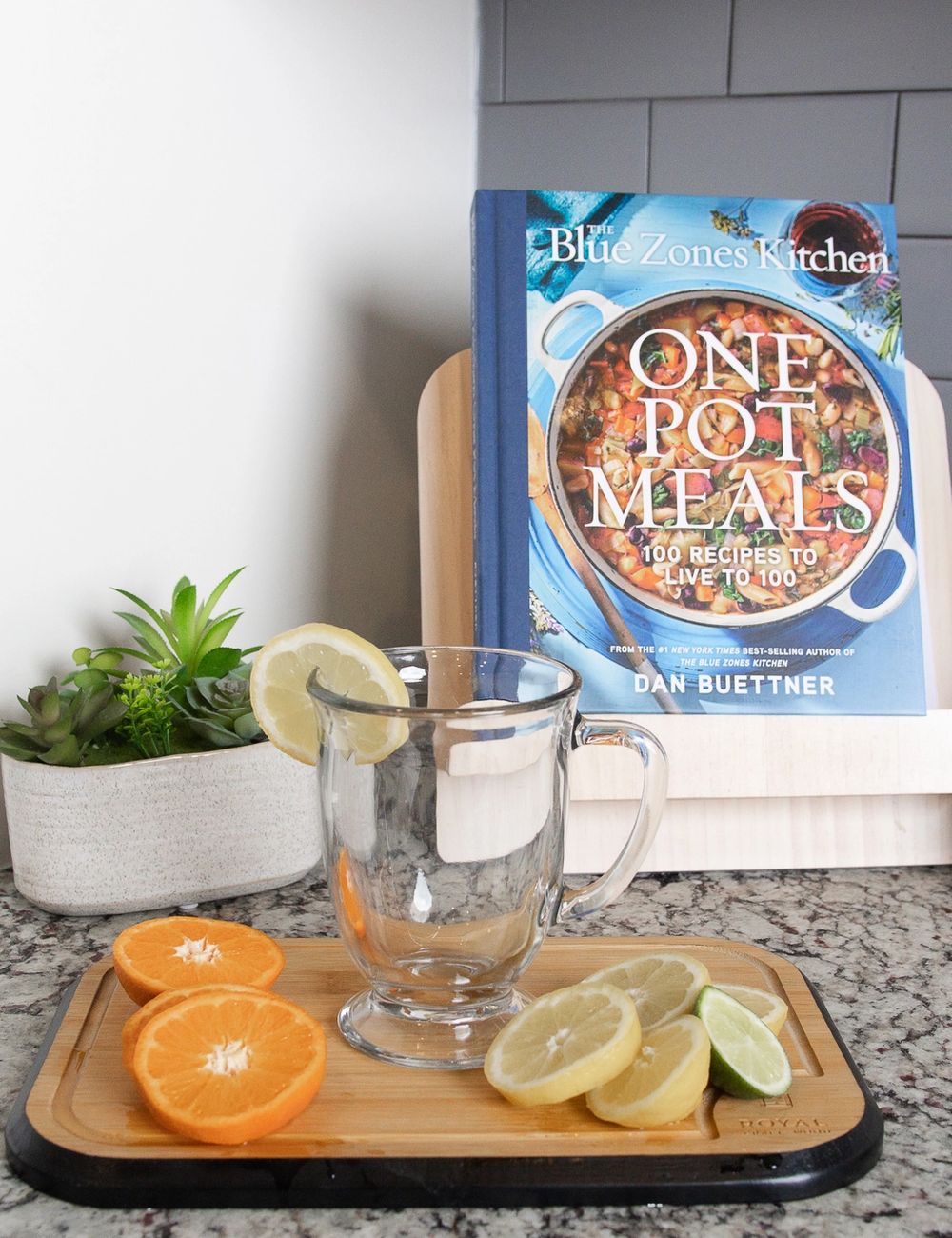 A clear glass and matching tall carafe of citrus-cooled infused water on a wooden tray, lemon and orange wedges arranged beside the glass, kitchen counter at daylight.