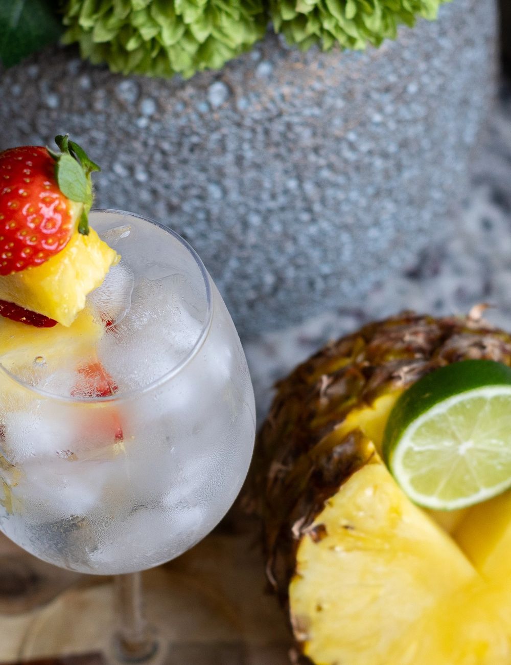 Tall wine glass of sparkling water over ice, strawberry-and-pineapple skewer balanced across the rim, lime wedge and pineapple wedge styled at the base, dark tile backdrop.