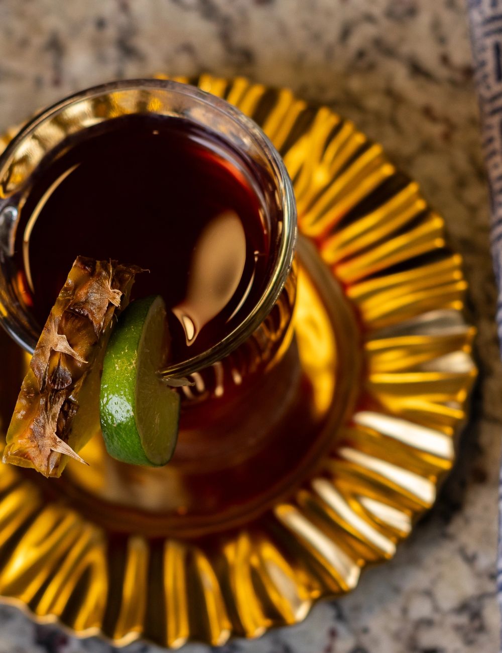 Overhead shot of a faceted glass mug holding deep amber tea, garnished with a cinnamon stick and a lime wedge, on a gold-rayed scalloped saucer.