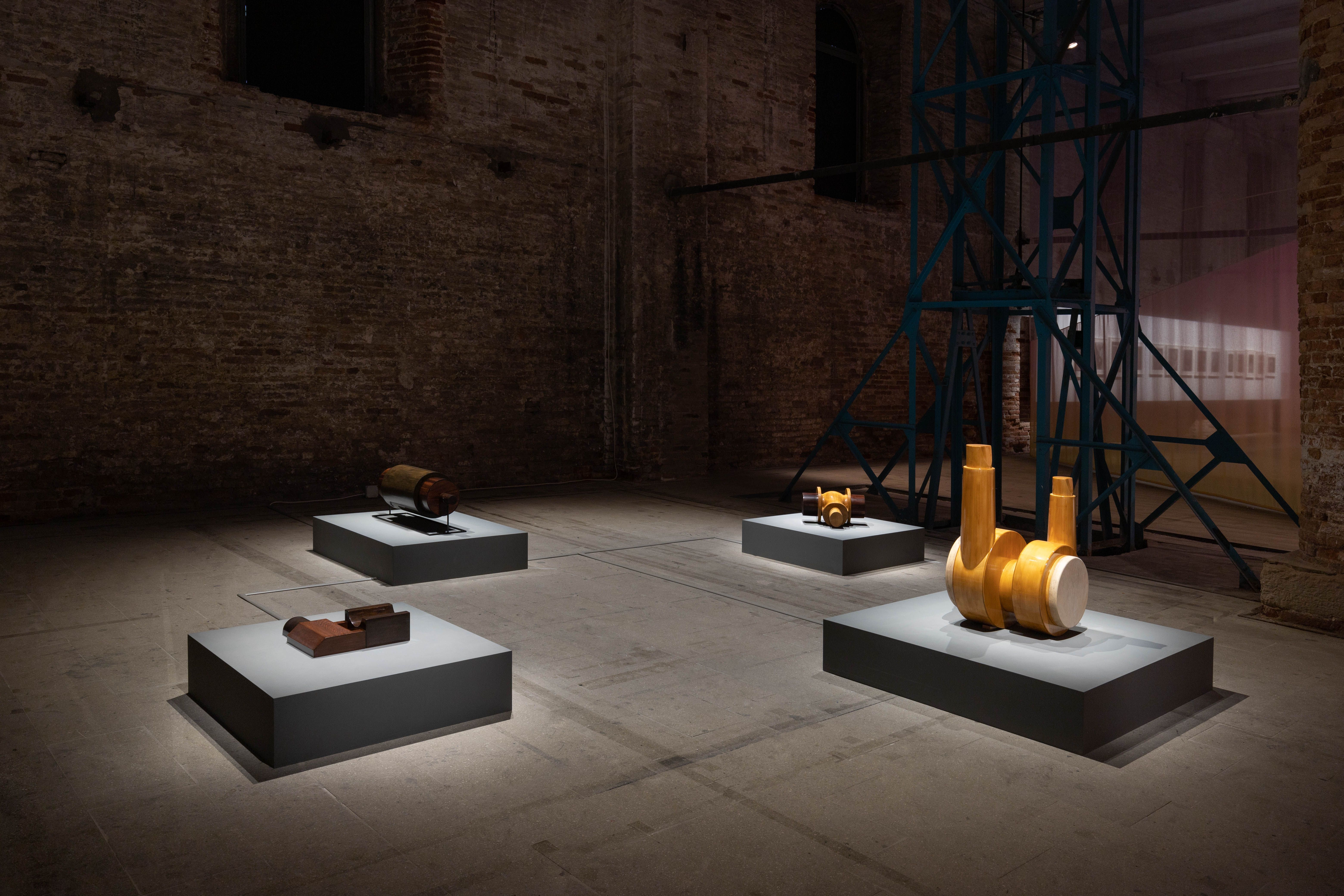 An installation view of Dora Budor's Autophones at the Venice Biennale. Four low gray plinths, each holding a different wood and metal sculptural component, are spread across a dimly lit, industrial brick hall. To the right, a tall teal metal structure and a translucent pink curtain stand in the background.