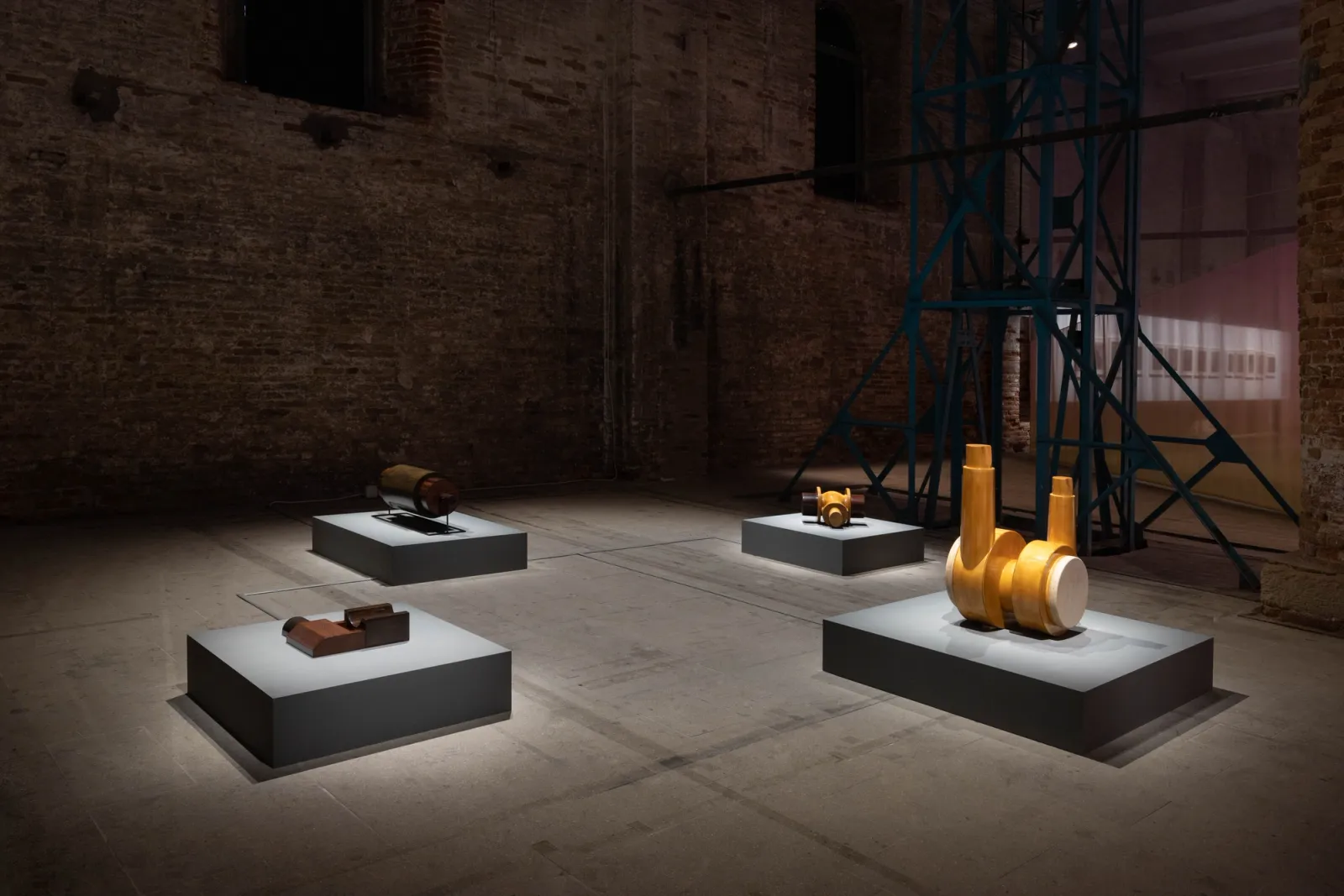 An installation view of Dora Budor's Autophones at the Venice Biennale. Four low gray plinths, each holding a different wood and metal sculptural component, are spread across a dimly lit, industrial brick hall. To the right, a tall teal metal structure and a translucent pink curtain stand in the background.