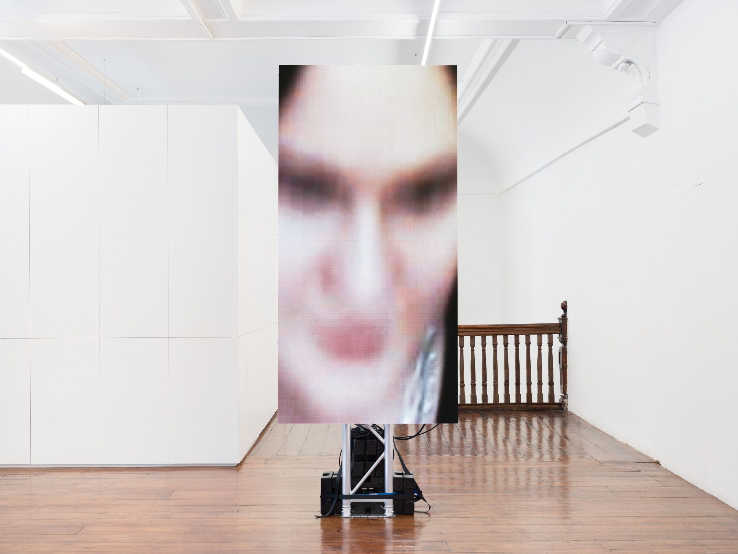 A vertical LED screen in a white gallery displays a blurred, pixelated close-up of a face.  The screen stands on a metal truss base atop polished wood floors near a wooden banister.