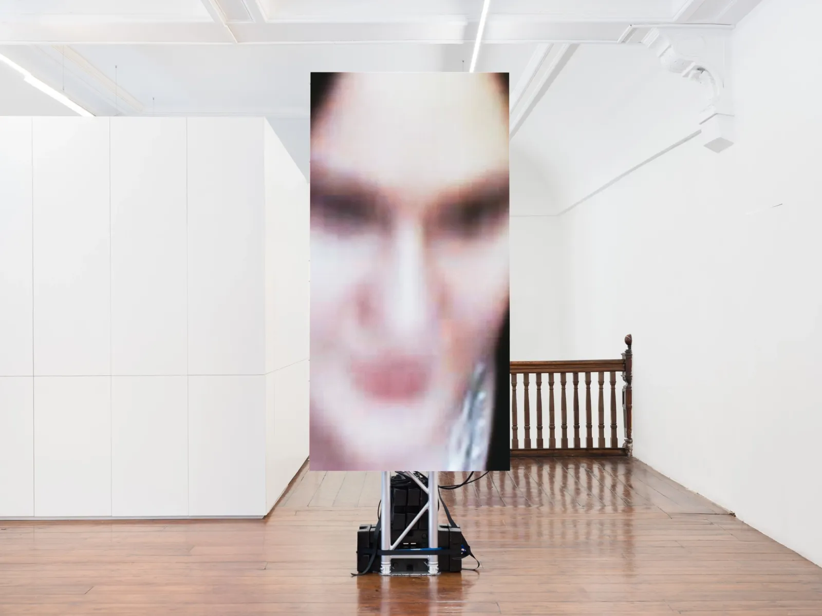 A vertical LED screen in a white gallery displays a blurred, pixelated close-up of a face. The screen stands on a metal truss base atop polished wood floors near a wooden banister.