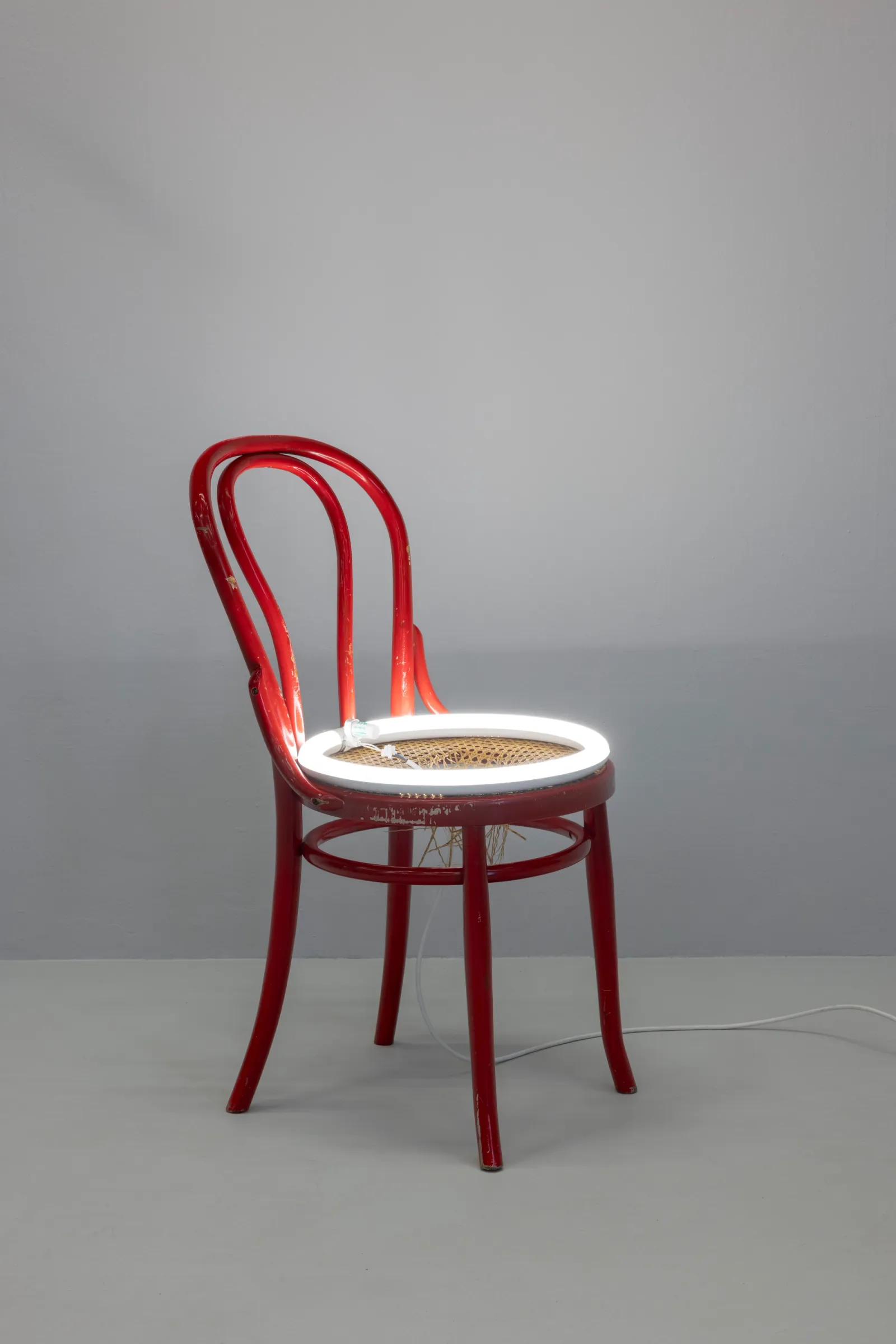 La scultura di Davide Stucchi: una sedia in legno rossa in stile Thonet con una luce al neon bianca circolare che illumina la seduta intrecciata.