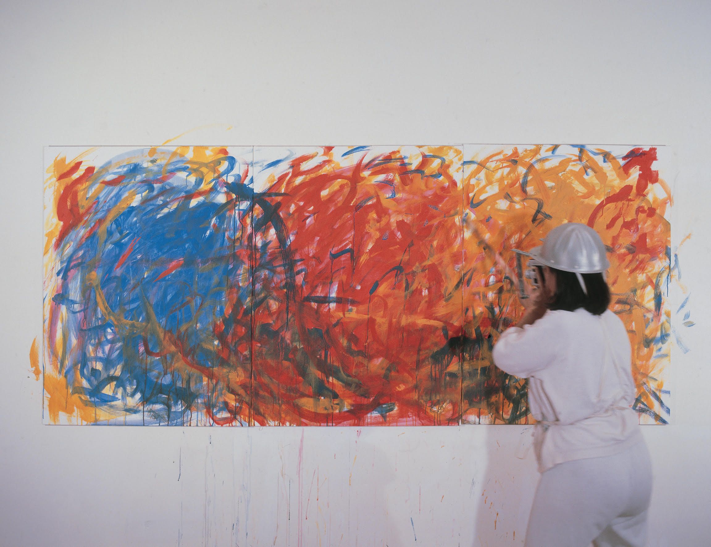 Margaret Raspé wearing a silver camera helmet stands before a large, three-panel abstract painting of energetic blue, red, and orange brushstrokes.