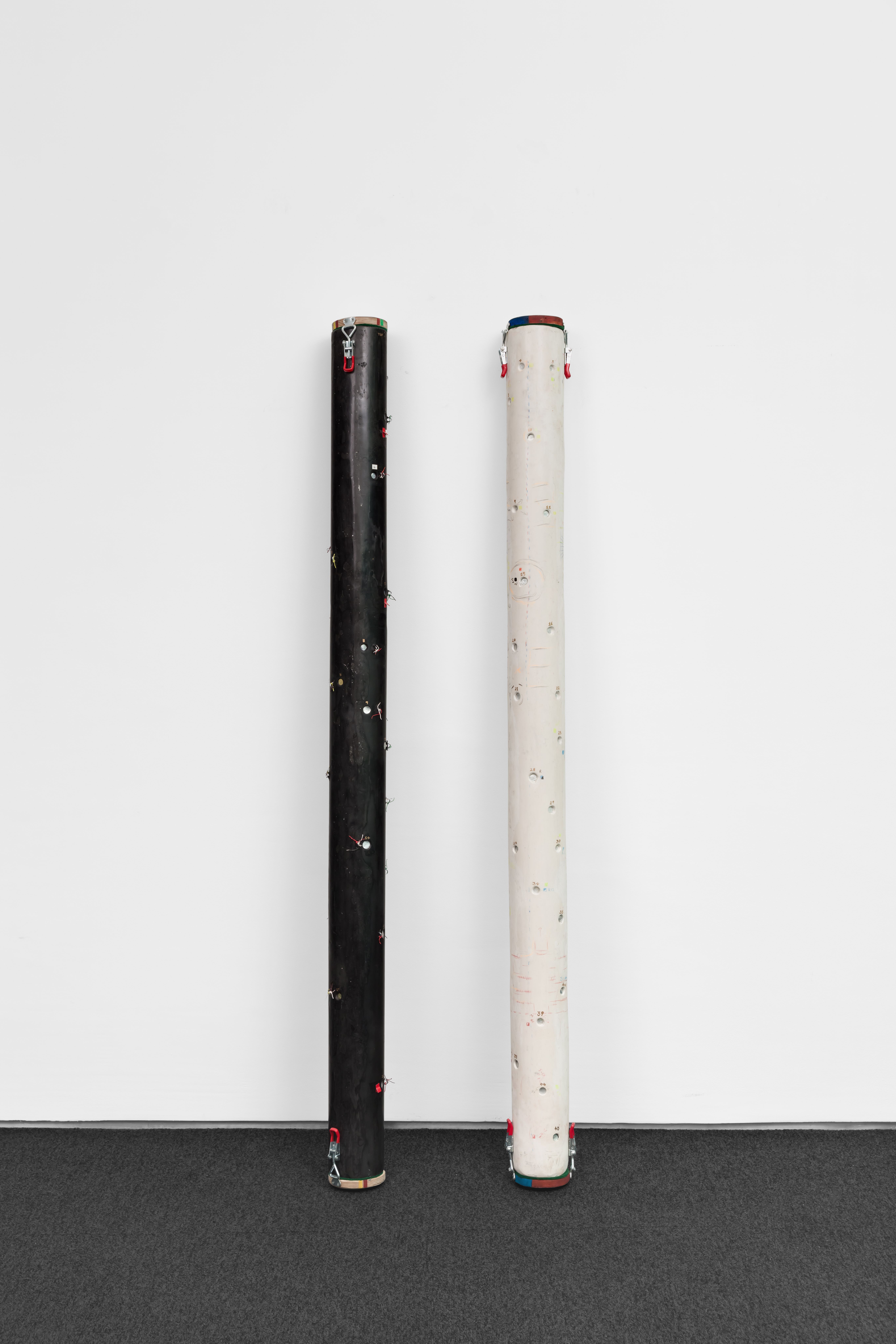 Two tall, slender cylindrical cameras—one black and one white—standing upright against a white wall. Both feature metallic latches and small lens apertures along their length.