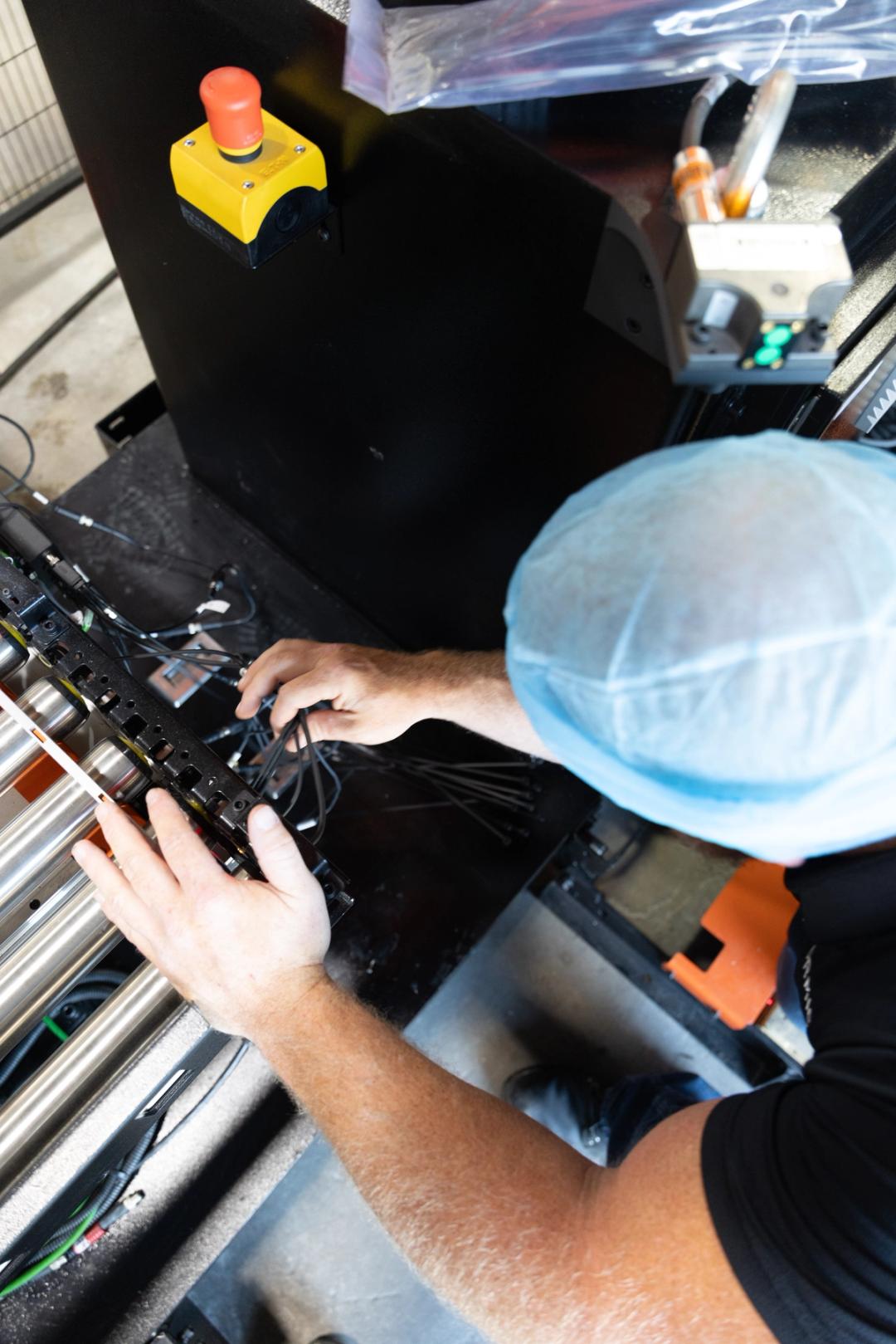 Formic field service technician in a hairnet connecting wiring on a robotic cell conveyor next to an emergency stop button