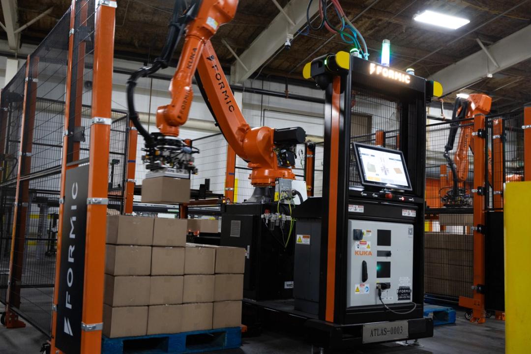 Formic Atlas robotic palletizer with a KUKA arm stacking cases onto a pallet inside a fenced cell with a touchscreen HMI