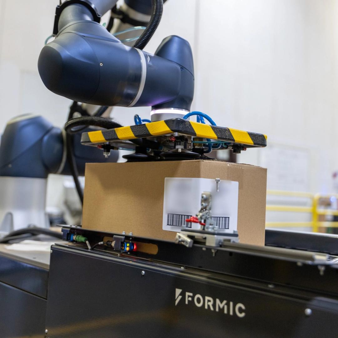 Formic cobot palletizing system using a vacuum gripper to lift a labeled corrugated shipping box off a Formic-branded conveyor during end-of-line automation.
