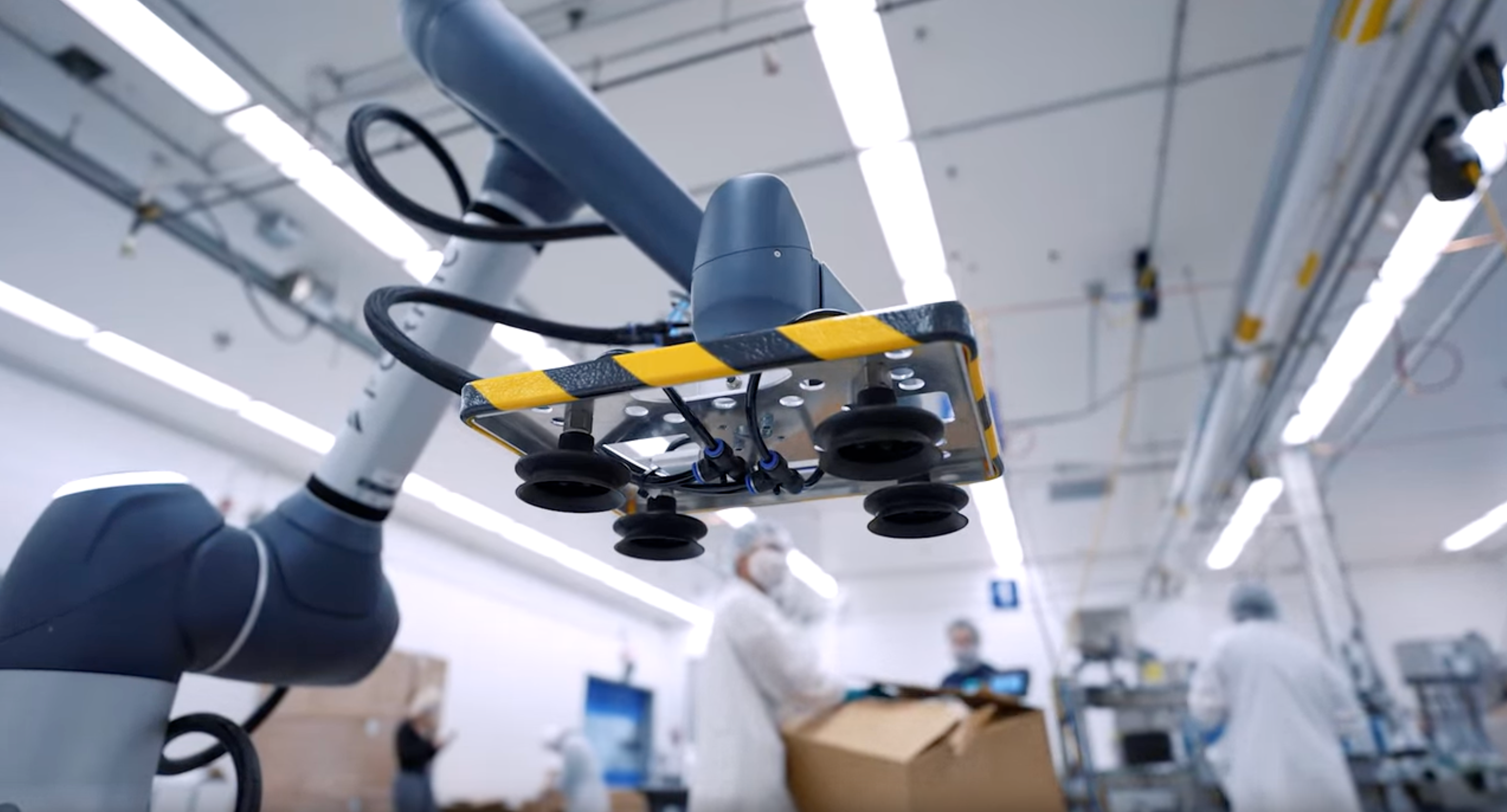 Close-up of a Formic collaborative robot arm with a yellow and black striped vacuum gripper and suction cups, positioned above a conveyor in a manufacturing facility.