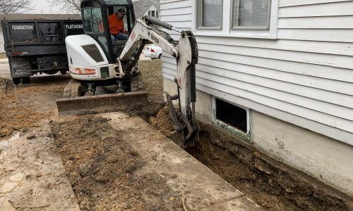 Photo of a small white excavator digging alongside a basement wall before the installation of Gorilla Wall Braces