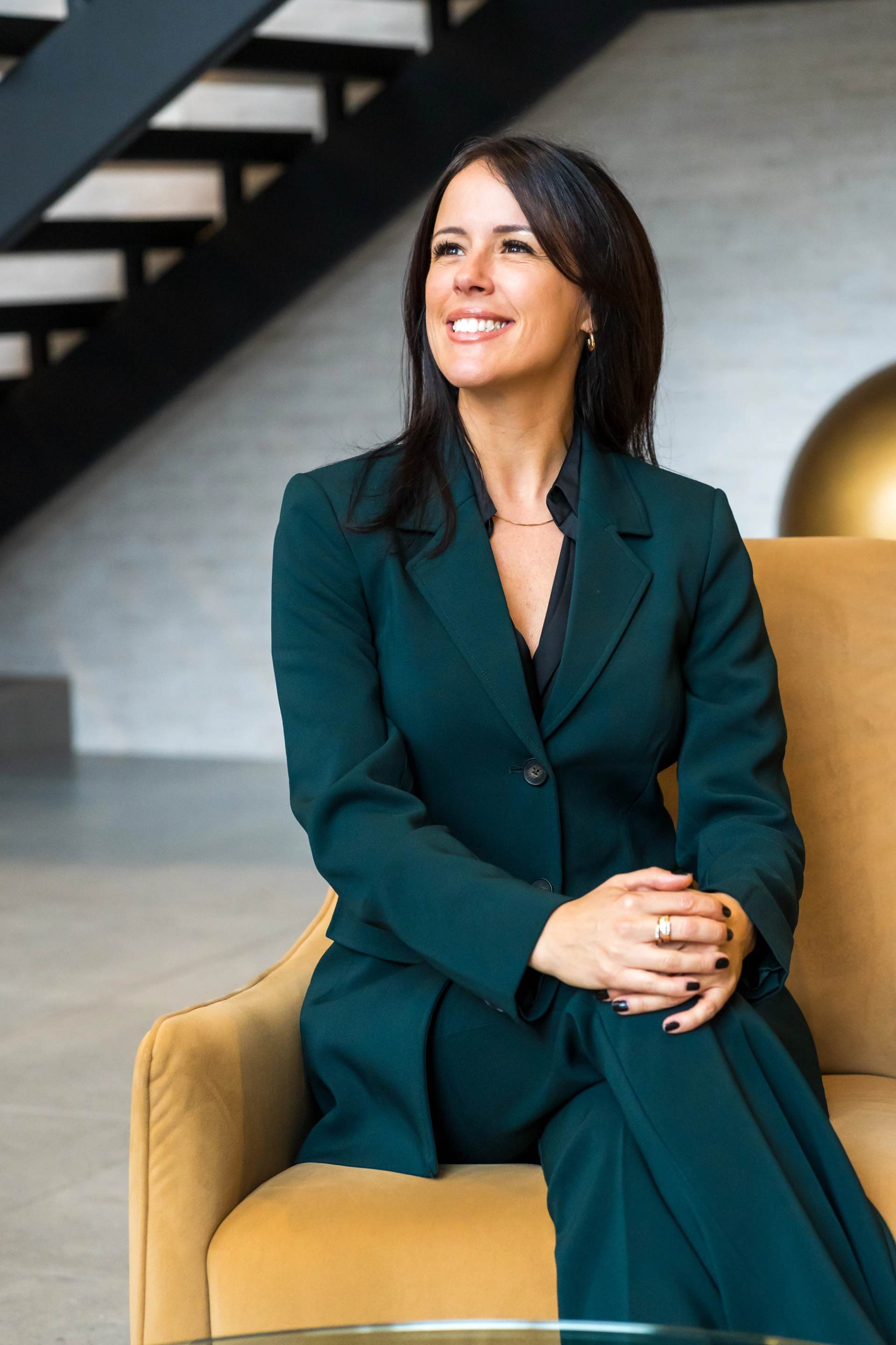 Femme en blazer vert assise dans un fauteuil jaune, escalier en arriere-plan