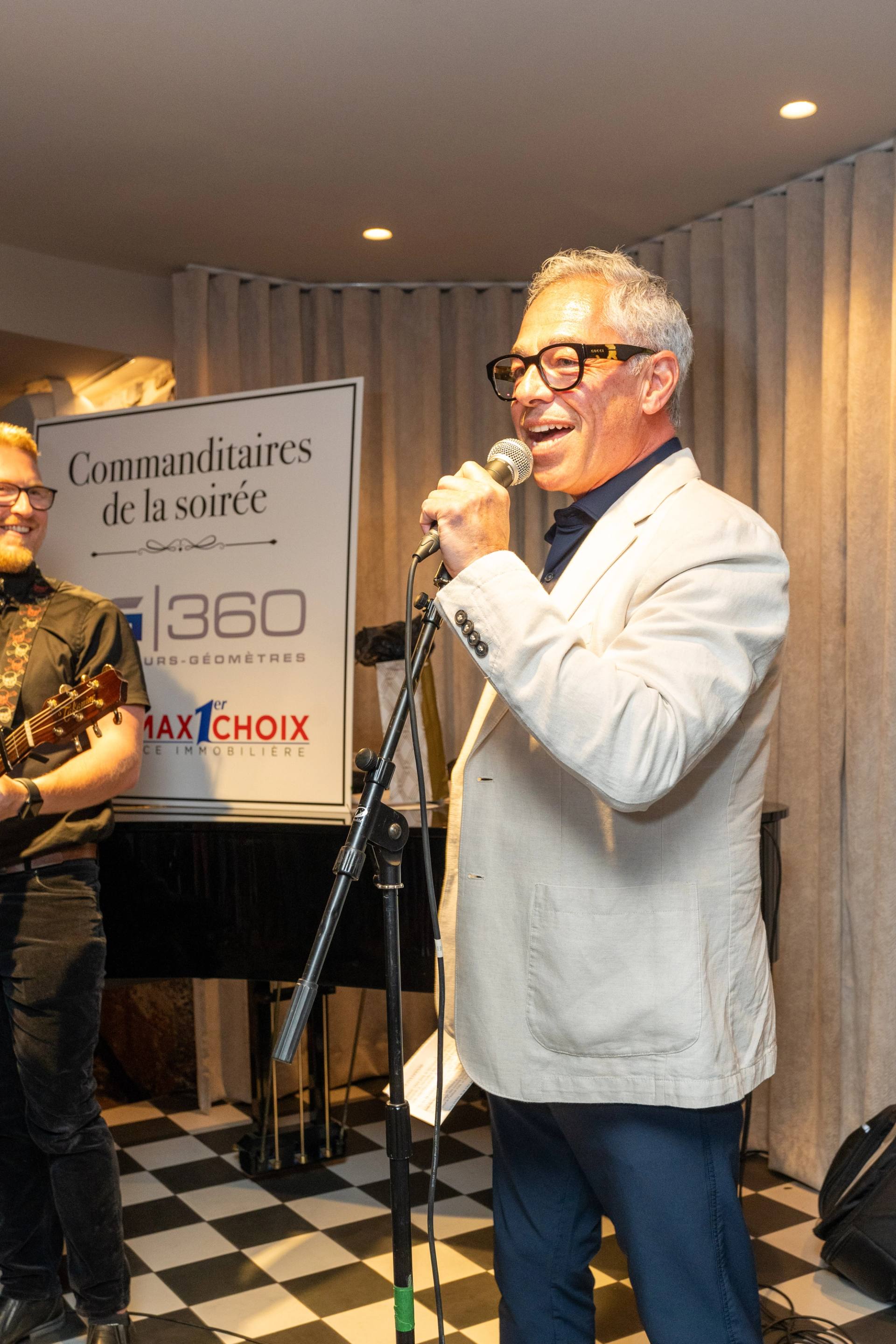 Animateur en veston blanc au micro devant le panneau des commanditaires de la soiree