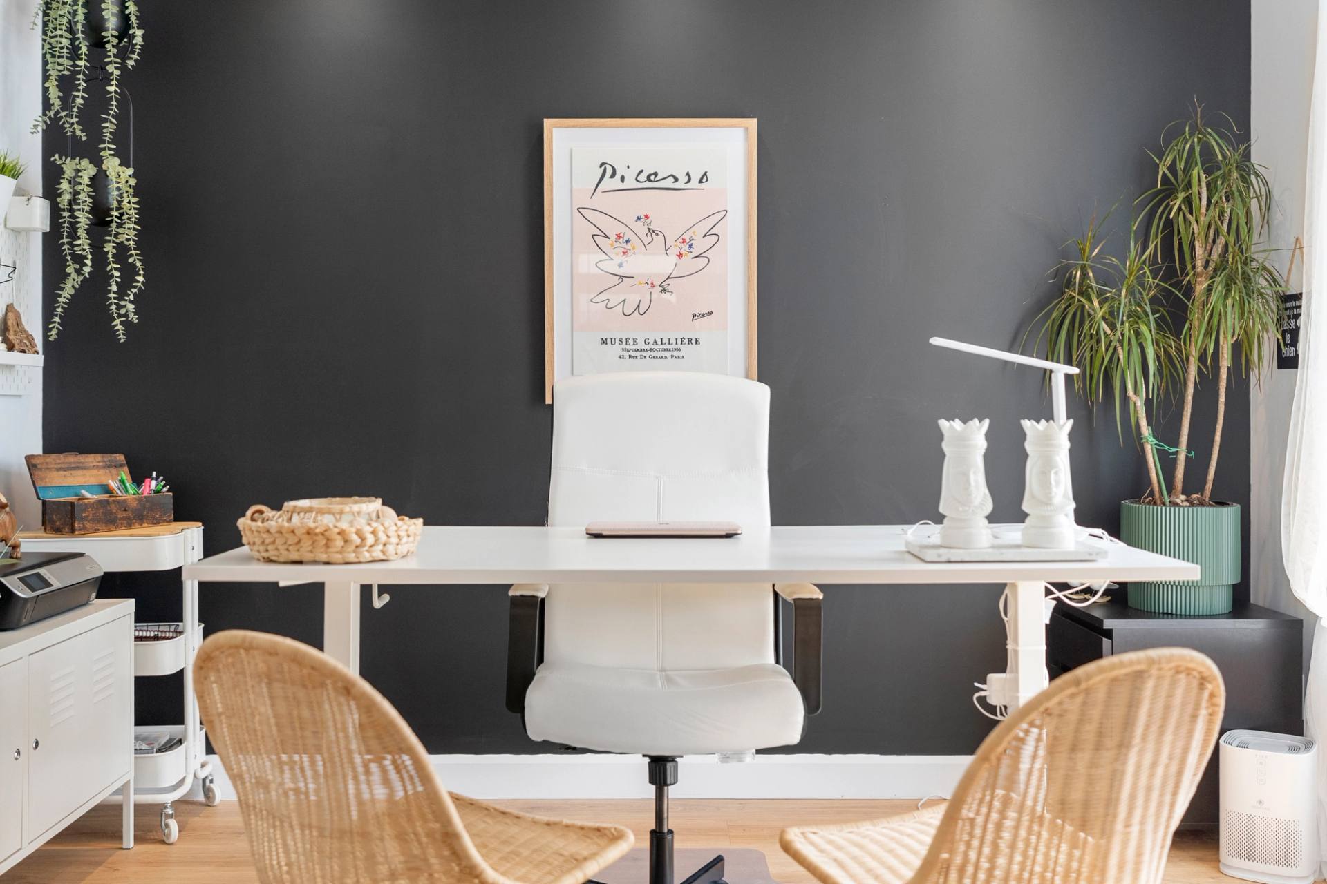 Coin bureau devant un mur noir, chaise blanche, fauteuils de rotin et plantes suspendues