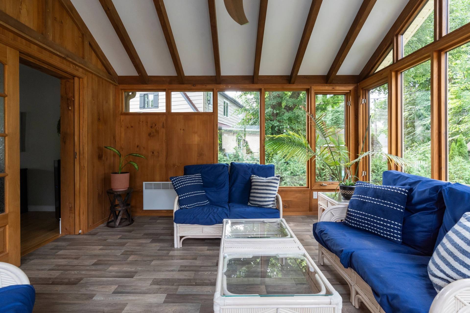 Solarium lambrissé de bois avec causeuses bleues, grandes fenêtres sur la verdure et embrasure sombre à gauche