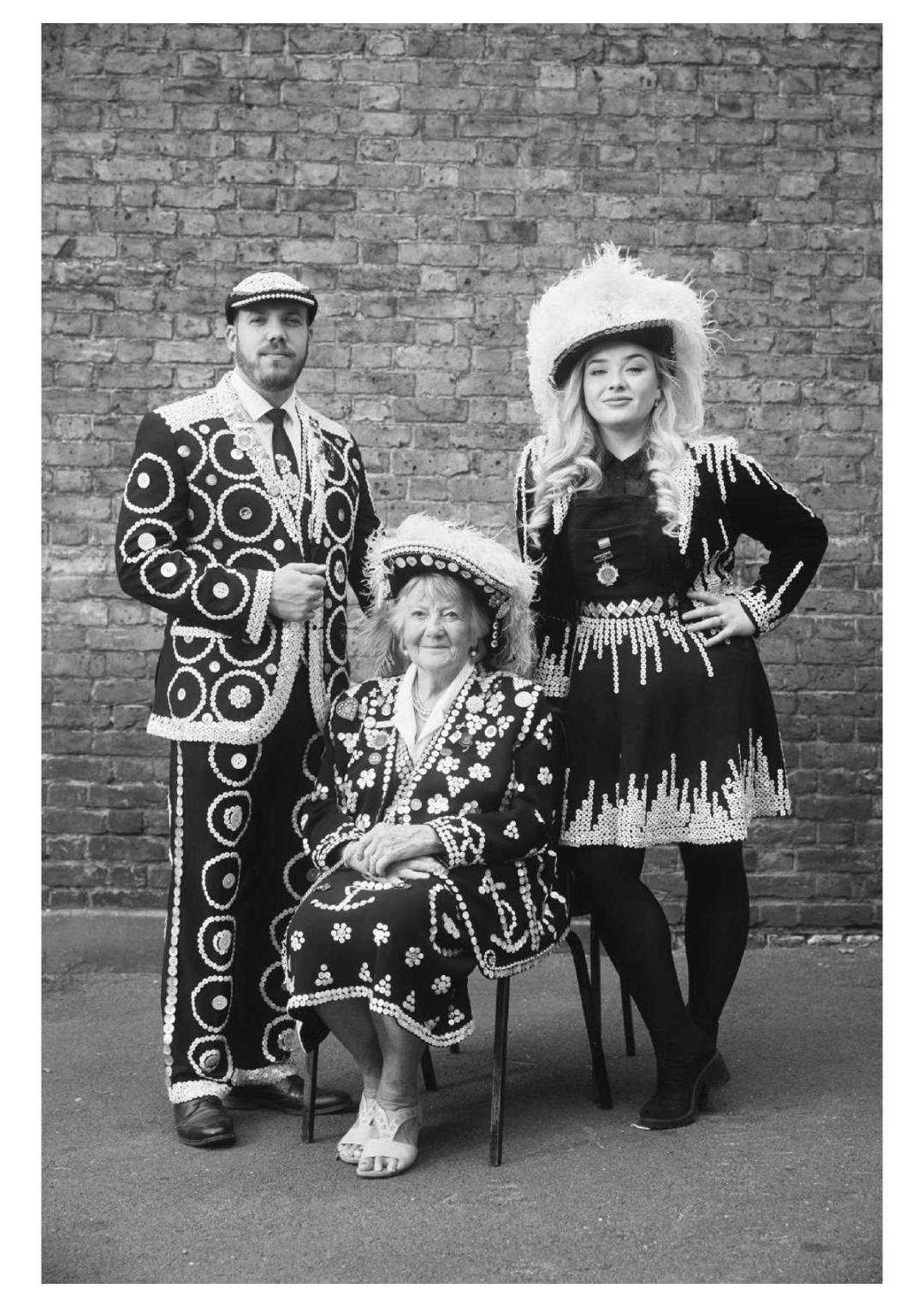 Black and white portrait of three Pearly Kings and Queens in ornate button-covered suits and feathered hats, against a brick wall.