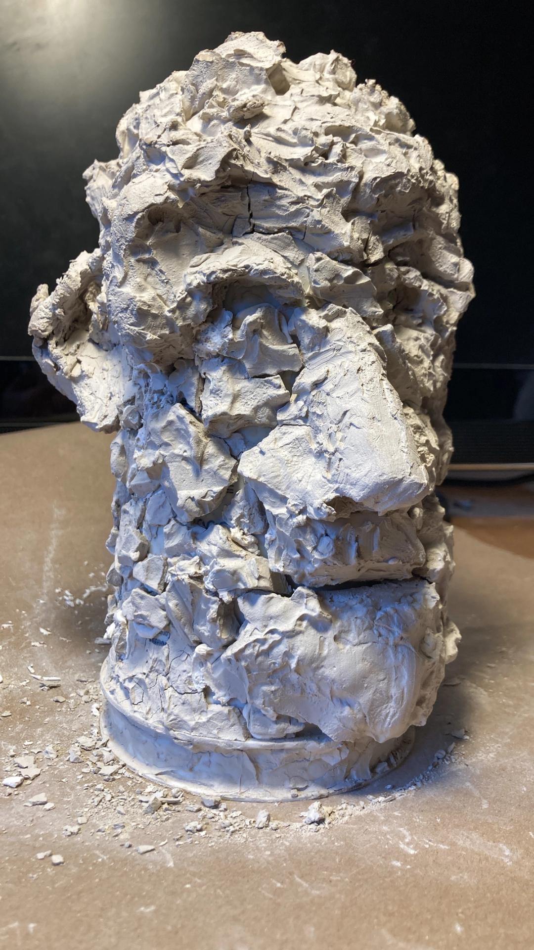 Raw unfired clay sculpture with a rough, cracked, rock-like surface on a wheel base, with clay fragments scattered around it.