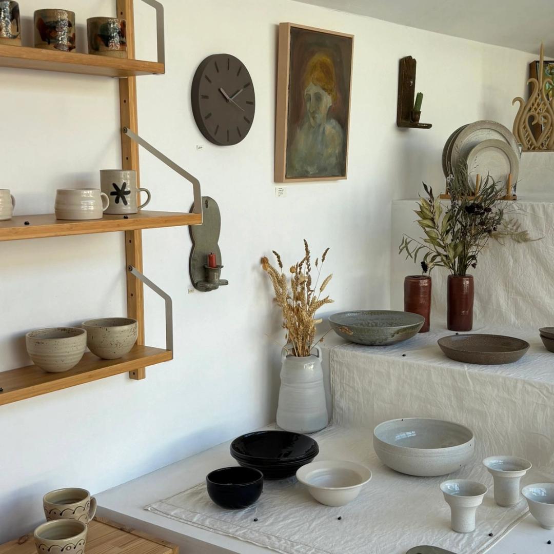Pottery shop display: shelves of handmade ceramic mugs and bowls, a wall clock, painted portrait, vases with dried wheat.
