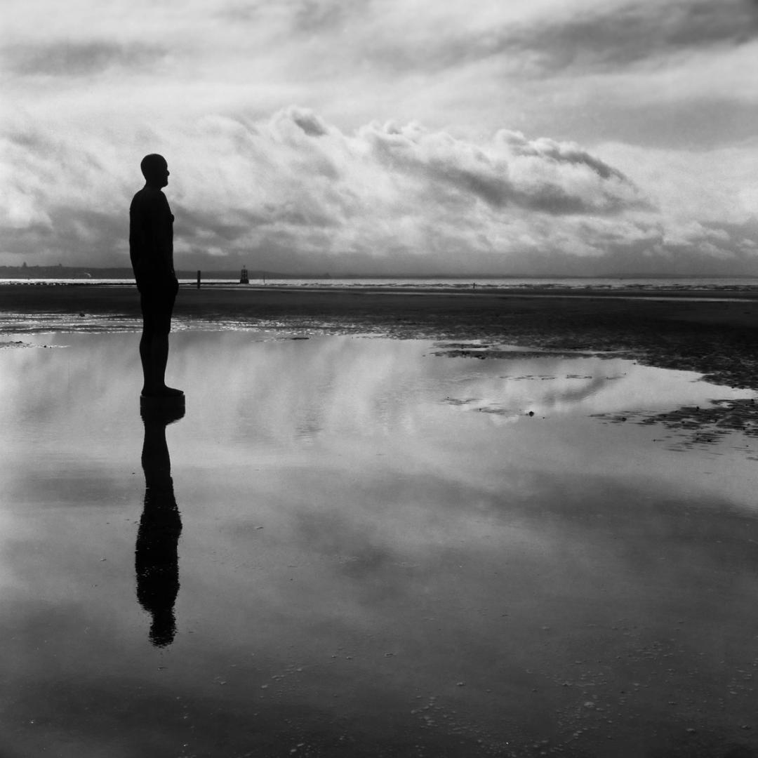 Black and white photo of an Antony Gormley statue reflected in the shallow water on the Beach at Crosby, Liverpool