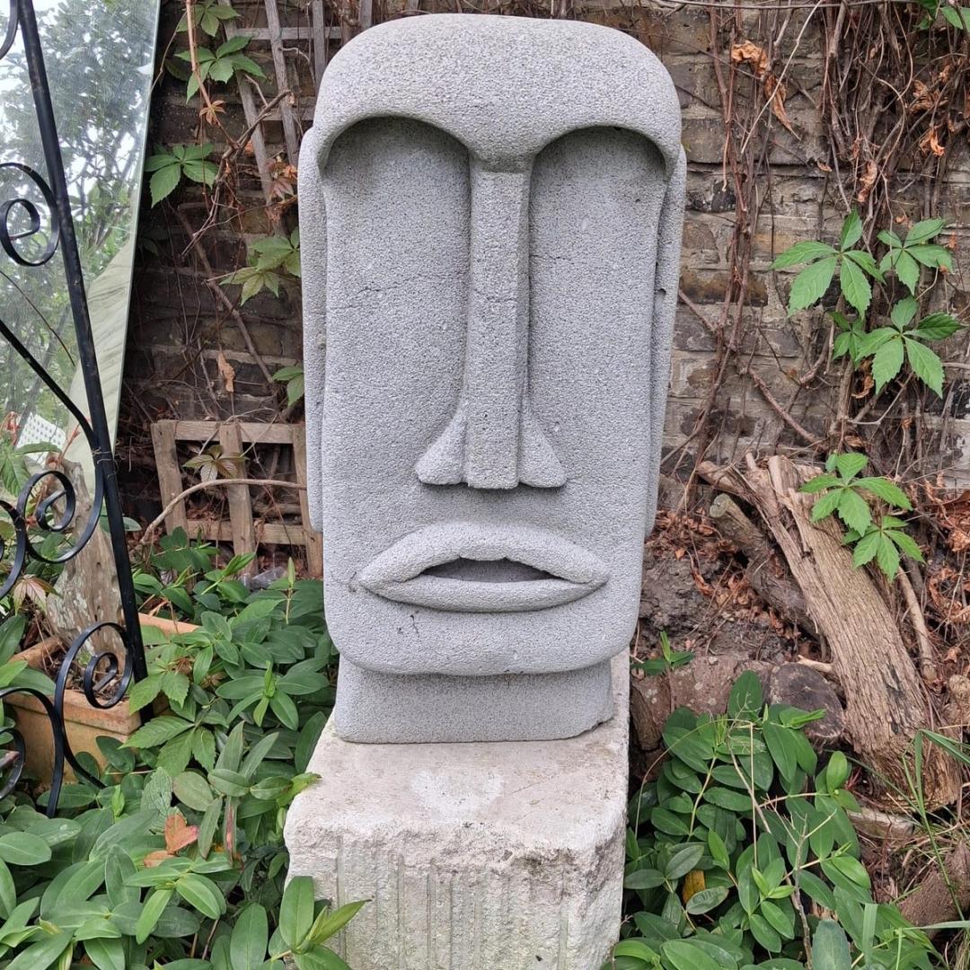 Grey stone garden sculpture of a Moai-style face on a plinth, set against a brick wall with climbing ivy and dried vines.