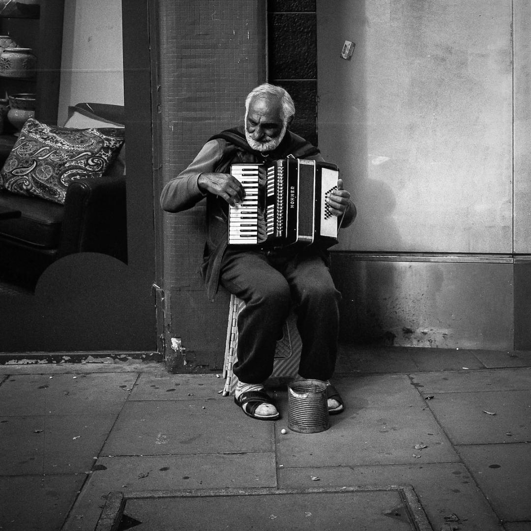 Black and white photo of an elderly man busking on a pavement, playing an accordion with a tin can at his feet.