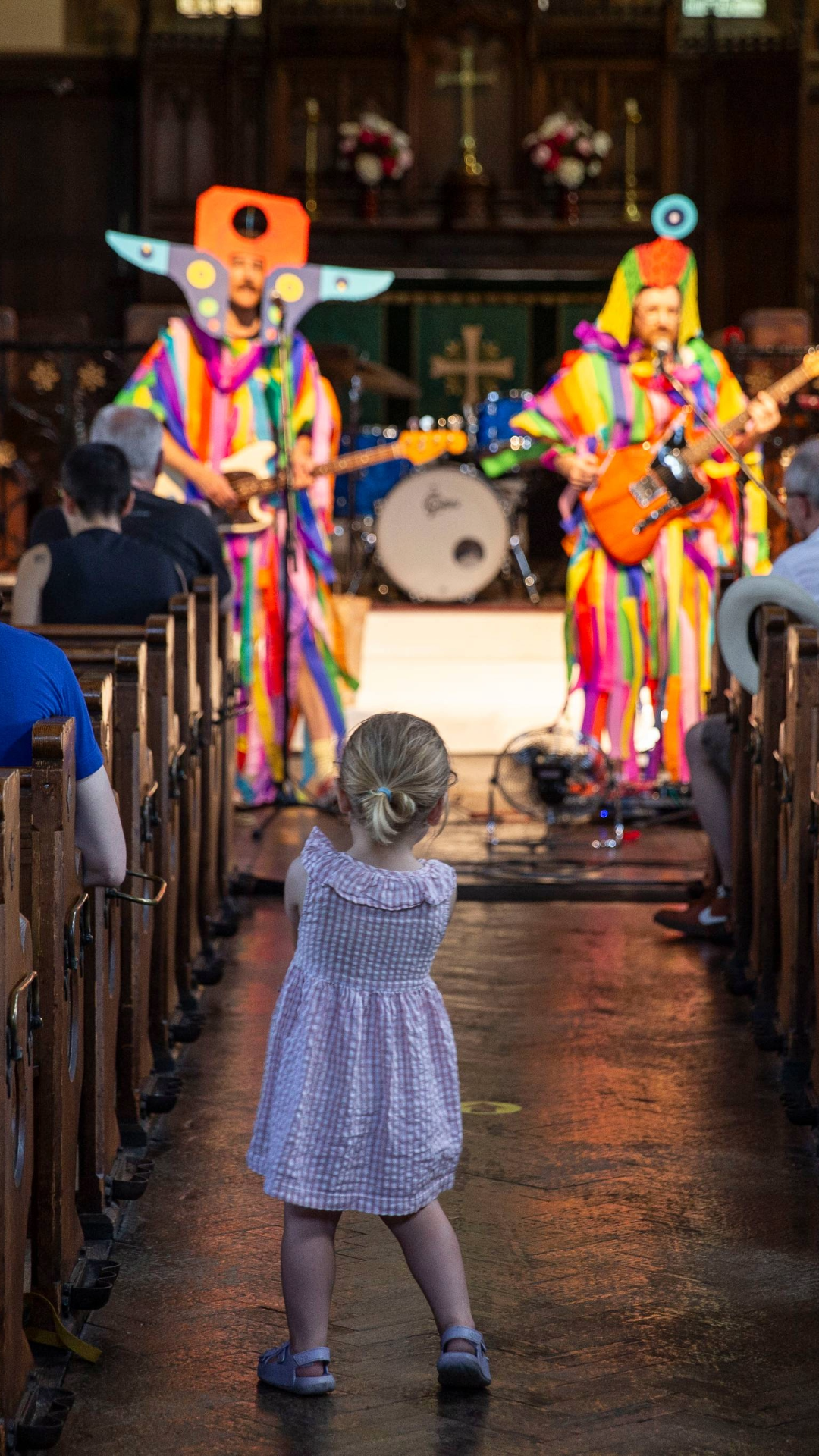 A toddler watches two musicians in rainbow costumes performing in a church aisle, as the congregation looks on from the pews.