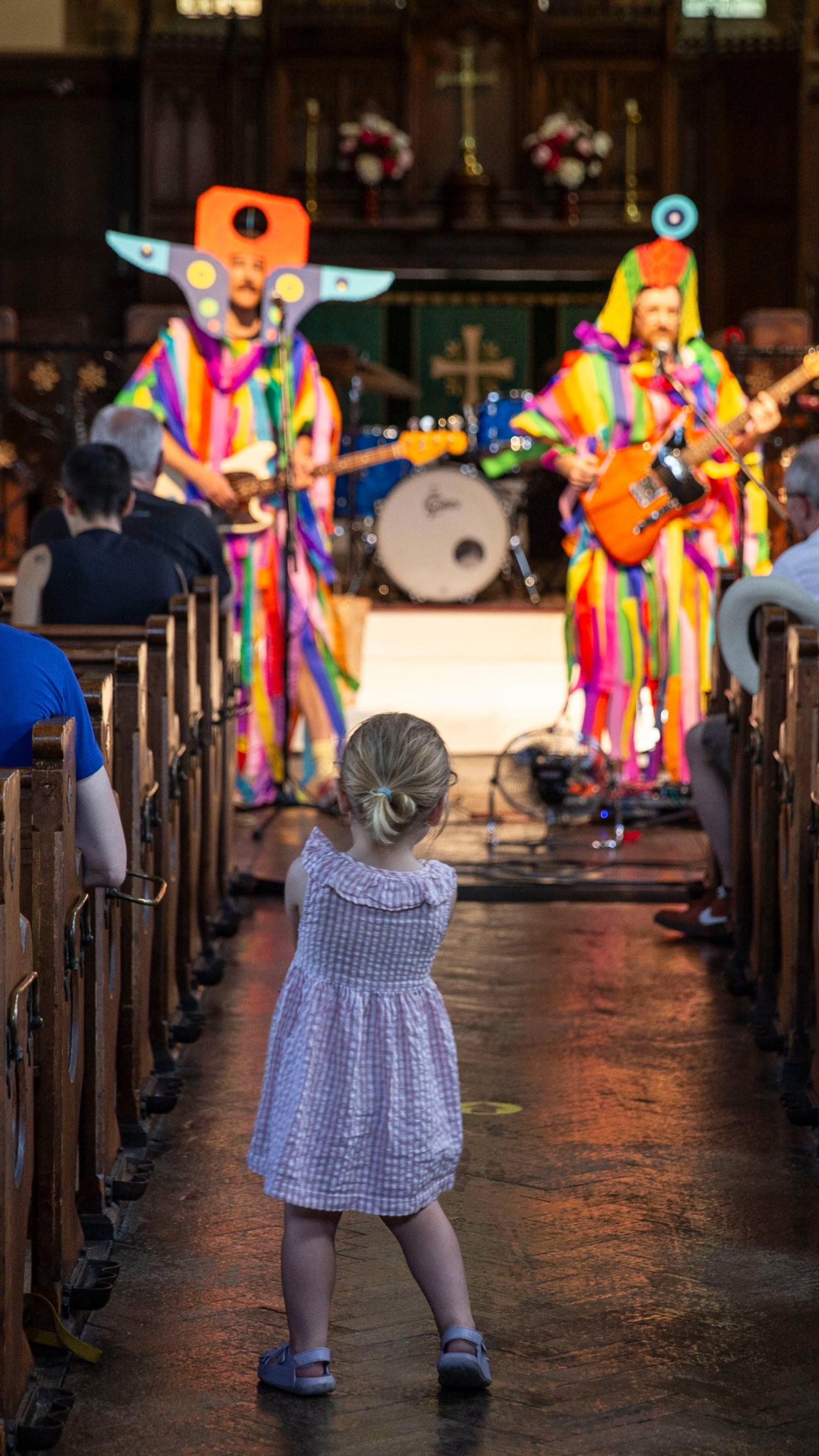A toddler watches two musicians in rainbow costumes performing in a church aisle, as the congregation looks on from the pews.