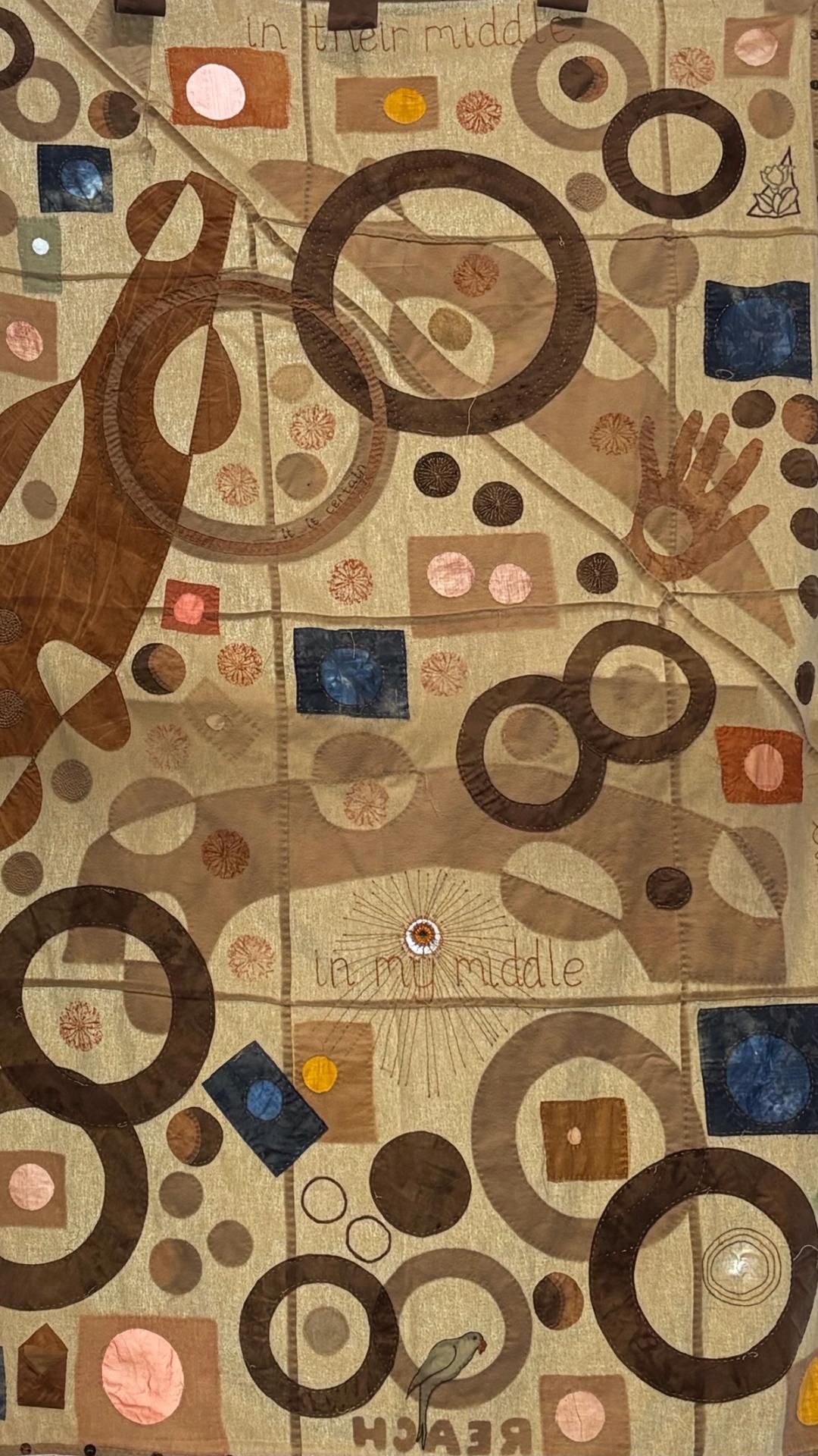 Large textile quilt with appliquéd circles, squares and embroidered text reading "in their middle" and "in my middle", in earth tones.