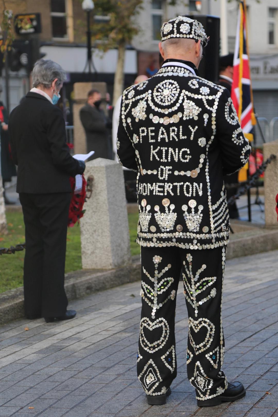The Pearly King of Homerton seen from behind in a button-covered suit at an outdoor ceremony, a Union Jack flag visible nearby.