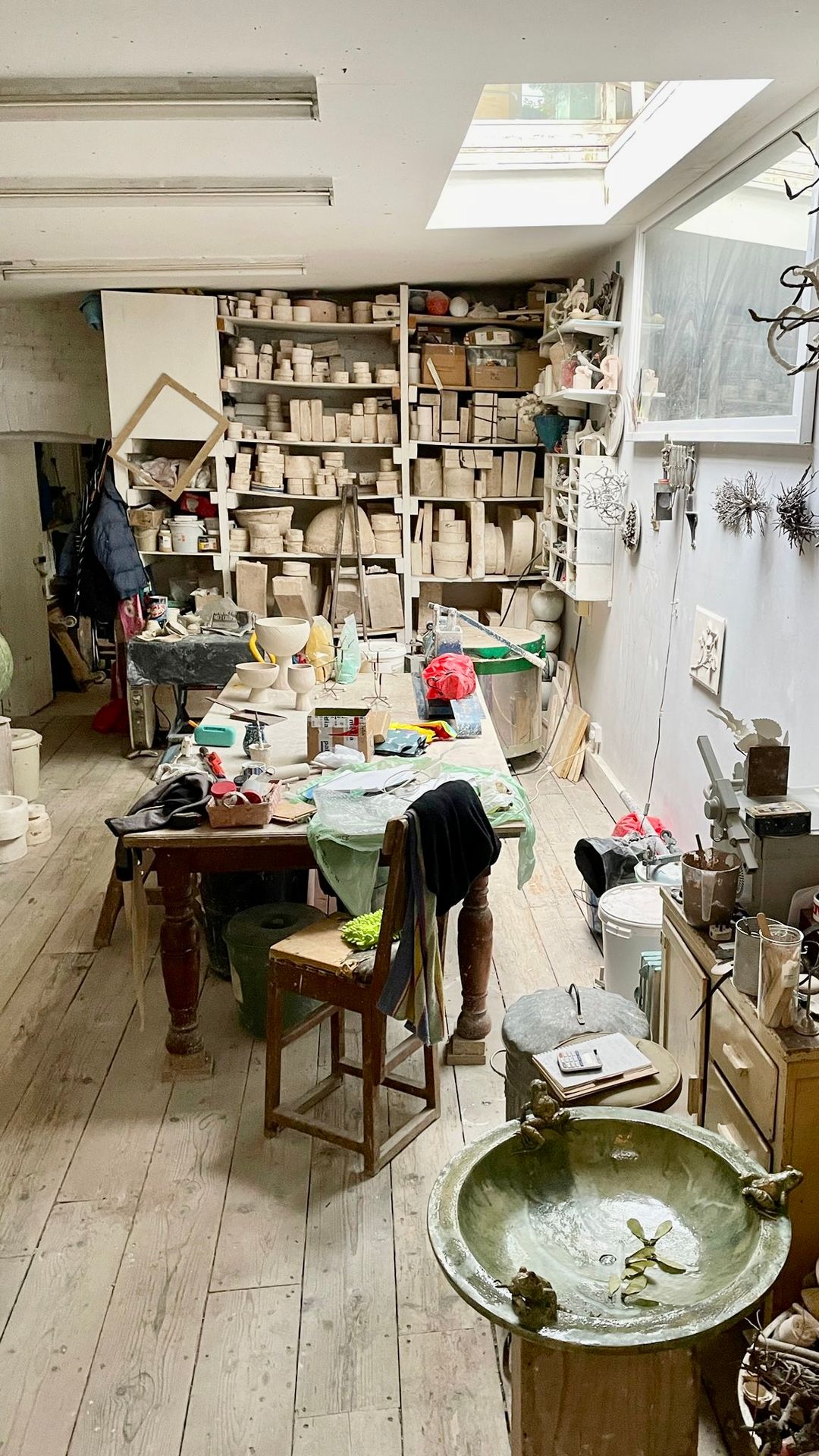 A busy ceramic artist's studio with shelves of clay works, a cluttered workbench, skylight, and a green glazed bowl in the foreground.