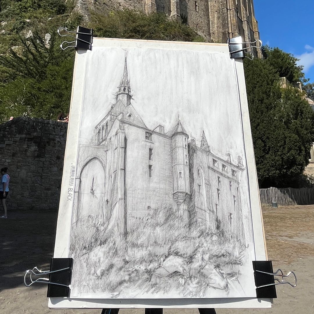 Charcoal sketch of a Gothic castle with spire and turrets, clipped to a drawing board, with the real structure visible behind.