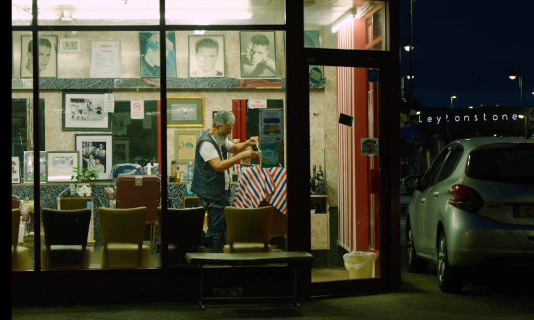 Photograph of a barbers shop from outside at night time, with a barber cutting someone's hair