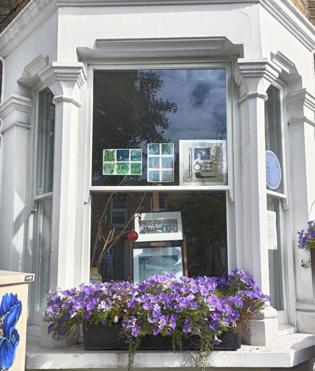 Artworks displayed in a bay window of a white Victorian terraced house, with overflowing purple petunias in the window box below.