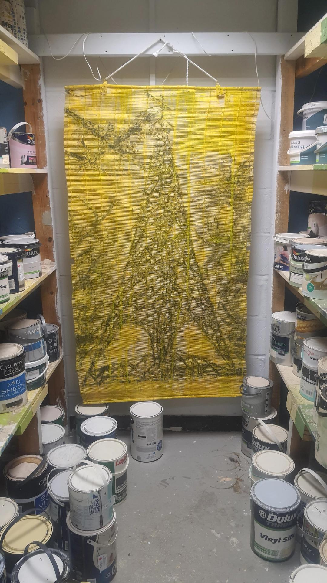 A yellow bamboo blind painted with a dark pylon and foliage, hung in a paint shop surrounded by tins of Dulux and Crown paint.
