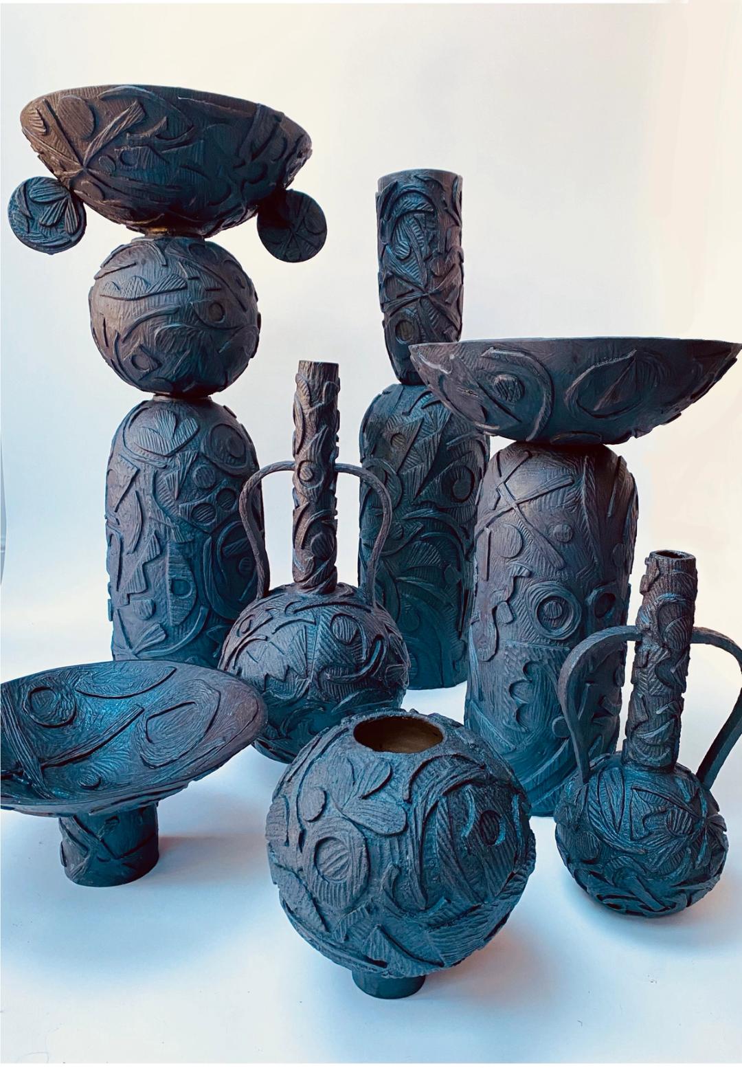 Collection of dark hand-built ceramic vessels with deeply carved botanical and abstract surface patterns, in various sculptural forms.